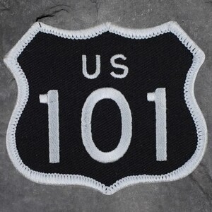 May include: Black and white patch in the shape of a US highway sign. The patch features the text "US 101" in white, with a white border. The background is black.
