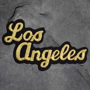 May include: A black embroidered patch with the words "Los Angeles" in gold script. The patch has a black border and is set against a gray, textured background. The lettering is in a cursive style.