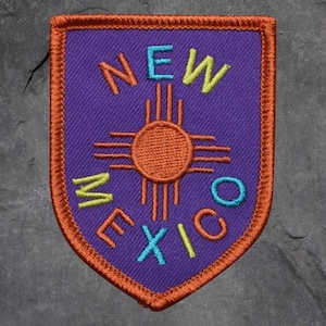 Peut inclure: Écusson brodé en forme de bouclier violet avec une bordure orange. Le patch présente le texte "NEW MEXICO" en différentes couleurs, avec un symbole de soleil orange au centre.
