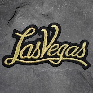 May include: Embroidered patch featuring the text "Las Vegas" in a cursive gold font, outlined in black. The patch is rectangular with rounded edges. The background is a dark gray, textured surface.