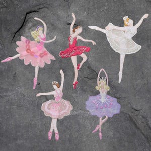 May include: Five fabric ballet dancer appliqués in various colours. Each dancer has a different tutu, including pink, red, and white, with sequin and glitter details. The dancers are posed in graceful ballet positions, with arms and legs extended.