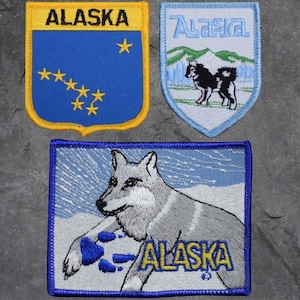Puede incluir: Tres parches bordados con la palabra "Alaska". El primer parche es azul con estrellas amarillas y un borde amarillo. El segundo parche es azul con una cordillera blanca y un perro blanco. El tercer parche es azul con un lobo blanco y la palabra "Alaska" en letras amarillas.