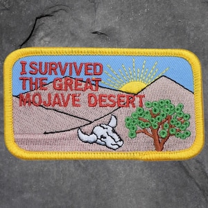 May include: Embroidered patch with a yellow border and the text "I Survived The Great Mojave Desert." The patch features a desert landscape with a sun, mountains, a tree, and a skull.