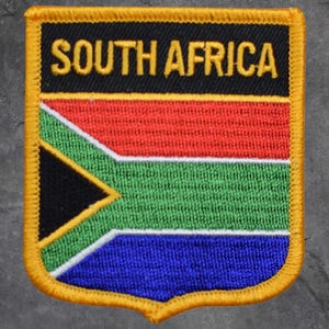 May include: Embroidered patch featuring the South African flag. The patch has a black top with "SOUTH AFRICA" in gold lettering. The flag design includes red, green, blue, and a black triangle with yellow trim. The patch is bordered with gold.