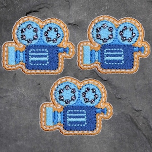 May include: Three embroidered patches in the shape of vintage movie cameras. The cameras are primarily blue with light blue and tan accents. The lenses have a floral design. The patches are outlined with tan stitching.