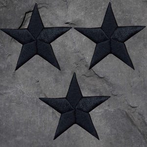 May include: Three black embroidered star patches on a grey, textured surface. Each star has five points and a detailed, stitched texture, creating a dimensional effect. The background is a mottled grey, resembling stone.