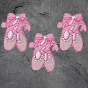 May include: Three pink ballet slipper patches with bows and rhinestone accents. The patches have a textured appearance and are embellished with silver-colored rhinestones. The background is a dark gray, textured surface.