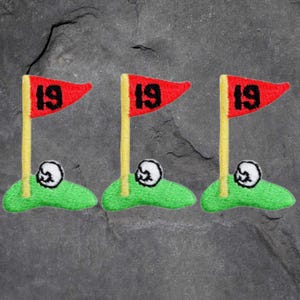 May include: Three embroidered golf patches. Each patch features a red flag with the number 19, a yellow flagpole, a white golf ball, and a green patch of grass. The patches are designed for apparel.