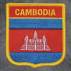 May include: Embroidered patch featuring the word "CAMBODIA" in yellow, above a blue stripe, a red field with a white and red temple design, and a blue stripe. The patch has a yellow border.