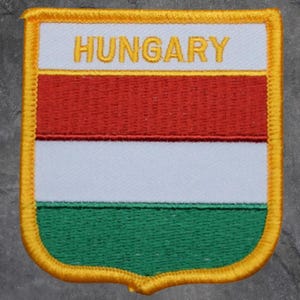 May include: Embroidered patch featuring the flag of Hungary. The patch has a shield shape with a gold border. The flag design includes horizontal stripes of red, white, and green, with the word "HUNGARY" in gold letters at the top.