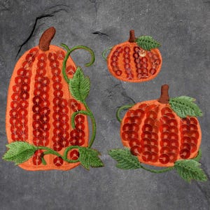 May include: Embroidered orange pumpkins with brown stalks and green leaves. The pumpkins are decorated with red sequins. One large pumpkin is tall and oval-shaped, with a green vine. Two smaller pumpkins are round.