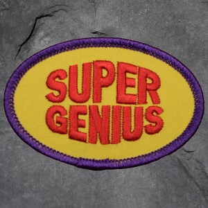 May include: Embroidered patch with the words "SUPER GENIUS" in red, bold letters on a yellow oval. The patch has a purple border with a stitched design. The background is a textured gray surface.
