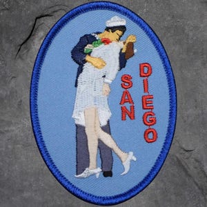 May include: Embroidered patch featuring a sailor embracing a woman, with the words "SAN DIEGO" in red. The patch has a blue oval border and depicts a classic romantic scene.