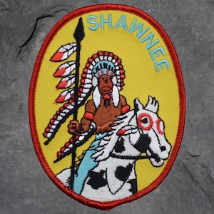 May include: Embroidered patch with a yellow background and a red oval border. The patch features a Native American figure riding a white horse with black spots. The word "SHAWNEE" is arched across the top in blue.