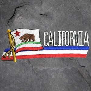 Könnte beinhalten: Gestickter Aufnäher mit dem Design der kalifornischen Staatsflagge. Die Flagge zeigt einen Braunbären, einen roten Stern und das Wort "CALIFORNIA" in Weiß. Der Aufnäher hat eine goldene Stange und blaue, weiße und rote Streifen.
