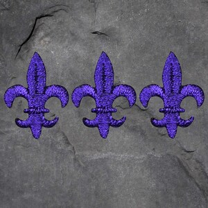 May include: Three embroidered purple fleur-de-lis patches. Each patch features a stylized design with three petals and a central vertical element. The patches are arranged horizontally against a dark gray, textured background.