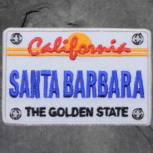 Peut inclure: Écusson brodé représentant une plaque d'immatriculation californienne. Le patch présente le mot "California" en script rouge au-dessus du texte bleu "SANTA BARBARA". En dessous, on peut lire "THE GOLDEN STATE". Le design comprend un soleil jaune et des rivets décoratifs.