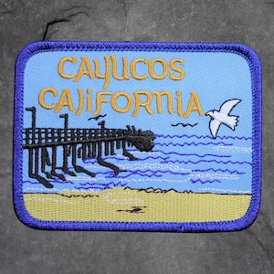 May include: Embroidered patch featuring a coastal scene with a pier, waves, and a seagull. The patch has the words "Cayucos California" in gold lettering against a light blue background, framed by a blue border.