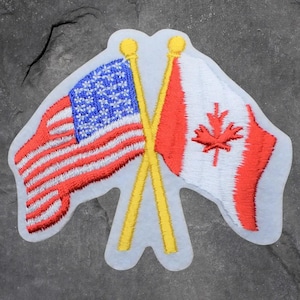 May include: Embroidered patch featuring the flags of the United States and Canada crossed. The American flag has red and white stripes with a blue field of white stars. The Canadian flag is red with a white maple leaf. The flags are on yellow poles.