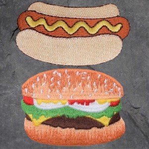 May include: Two embroidered felt food patches, a hot dog and a hamburger. The hot dog is yellow and brown with a white bun. The hamburger is brown, green, yellow, and white with a sesame seed bun.