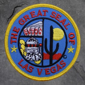 May include: Embroidered patch featuring "THE GREAT SEAL OF LAS VEGAS" in red lettering on a blue background. The design includes a cactus, a yellow sun, playing cards, a slot machine, dice, and two yellow stars, all framed by a gold border.