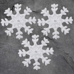 May include: Three white snowflake ornaments with intricate designs are arranged on a dark gray, textured surface. The snowflakes have detailed, symmetrical patterns, perfect for winter or holiday decorations. Each snowflake has six points.