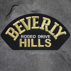 May include: Black embroidered patch with the words "BEVERLY HILLS" in gold thread. The text is arched across the top and bottom, with "RODEO DRIVE" in the center. The patch is in a fan shape.