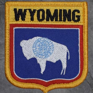 May include: Embroidered patch featuring the word "WYOMING" in black block letters above a blue shield. The shield displays a white bison with a blue circle on its side, outlined in white. The patch has a red and gold border.