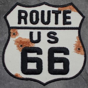 May include: Embroidered Route 66 patch in the shape of a road sign. The patch features the words "ROUTE US 66" in black, with a white background and brown distressed accents. The border is black.