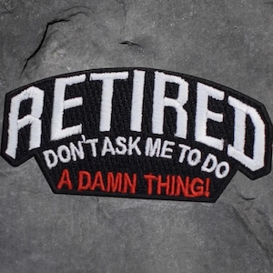 以下が含まれることがあります： 白いブロック体で「RETIRED」と書かれた黒い刺繍パッチ。その下に「DON'T ASK ME TO DO」と「A DAMN THING！」が赤で書かれています。パッチは黒い縁取りがあり、灰色の表面にあります。