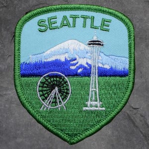 May include: Embroidered patch featuring Seattle landmarks. The design includes the Space Needle, a Ferris wheel, and a mountain range, with the city name in green letters against a light blue sky. The patch has a green border and a green grassy area.