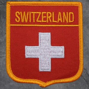 Peut inclure: Écusson rouge en forme de bouclier avec une croix blanche, l'emblème de la Suisse. Le mot "SWITZERLAND" est brodé en or au-dessus de la croix. Le patch a une bordure dorée.