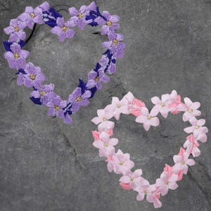 May include: Two floral heart-shaped hair accessories. One is lavender with gold accents and dark blue detailing. The other is pink with gold accents and light pink detailing. Both are on a gray stone surface.