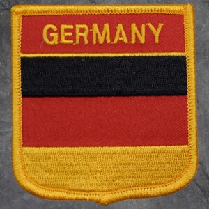 Peut inclure: Écusson rectangulaire avec le mot « GERMANY » en lettres dorées. En dessous, une bande noire et une bande rouge. L'écusson a une bordure dorée et une section inférieure jaune, ressemblant à un drapeau.