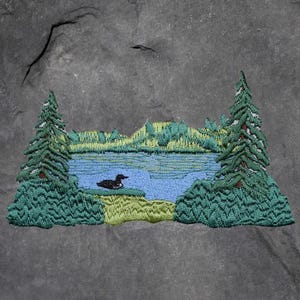 May include: Embroidered patch featuring a serene lake scene with a duck, evergreen trees, and a flying bird. The patch has a blue lake, green trees, and a gray background. The patch is likely for a jacket or bag.