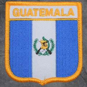May include: Embroidered patch featuring the flag of Guatemala. The design includes vertical blue and white stripes, with the Guatemalan coat of arms in the center. The word "GUATEMALA" is embroidered in gold at the top, with a gold border.