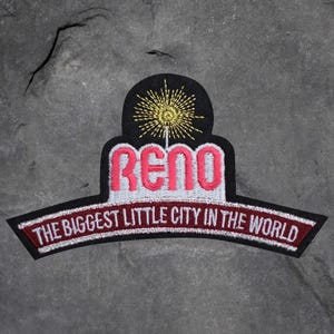 Könnte beinhalten: Gestickter Aufnäher mit dem Wort "RENO" in Rosa, über dem Satz "THE BIGGEST LITTLE CITY IN THE WORLD". Der Aufnäher hat einen schwarzen Hintergrund mit goldenen und weißen Akzenten.