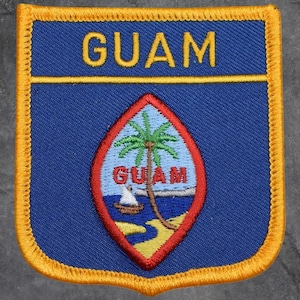 May include: Embroidered patch featuring the word "GUAM" in yellow above a shield design. The shield contains a palm tree, boat, and the word "GUAM". The patch has a blue background with a gold border.