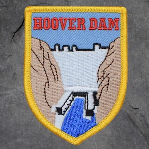 Könnte beinhalten: Gestickter Aufnäher mit dem Hoover-Staudamm. Der Aufnäher hat einen blauen Himmelshintergrund mit den Worten "HOOVER DAM" in Rot. Der Damm ist in Weiß und Schwarz dargestellt, mit braunen Klippen auf beiden Seiten. Der Aufnäher ist gelb umrandet.