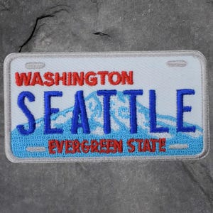 May include: Embroidered patch in the shape of a Washington state number plate. The patch features the words "WASHINGTON" in red, "SEATTLE" in blue, and "EVERGREEN STATE" in red, with a mountain graphic.