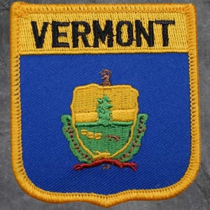 May include: A rectangular patch with the word "VERMONT" in black lettering above a blue background. The patch displays the state seal, with a yellow background, green tree, and red accents, all edged in yellow.