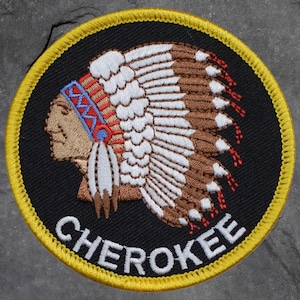 May include: Embroidered patch featuring a Native American chief in profile, wearing a feathered headdress. The patch is circular, with a black background, a yellow border, and the word "CHEROKEE" in white.