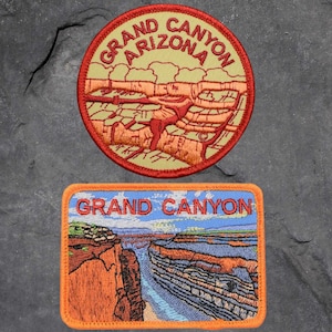 May include: Two embroidered patches featuring the Grand Canyon. The top patch is round and features the text "Grand Canyon Arizona" in red and white. The bottom patch is rectangular and features a colorful image of the Grand Canyon with the text "Grand Canyon" in red.