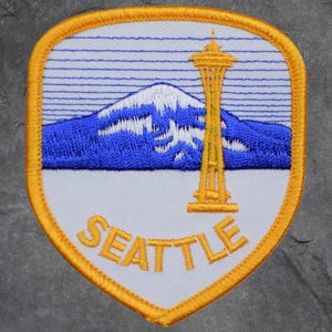 May include: Embroidered patch featuring a blue mountain range with a snow-capped peak and a yellow Space Needle. The word "SEATTLE" is embroidered in yellow. The patch has a white background and a yellow border.