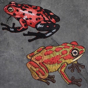 May include: Two embroidered frog patches. The first frog is red with black spots and has a black and white underside. The second frog is green with red spots and has a brown underside.