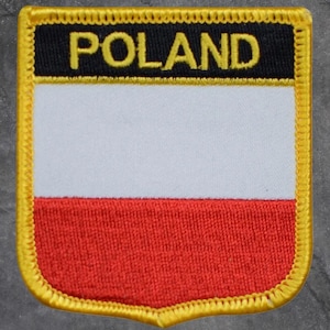 May include: Embroidered patch featuring the flag of Poland. The patch has a shield shape with a black top section with the word "POLAND" in yellow, a white middle section, and a red bottom section, all outlined in yellow.