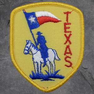 May include: Embroidered patch with a yellow background and a shield shape. The patch features a blue silhouette of a cowboy on a white horse holding a Texas flag. The word "TEXAS" is embroidered in red.