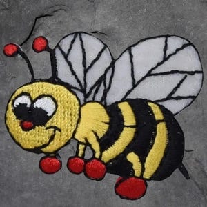May include: Embroidered patch featuring a cartoon bee with a smiling face. The bee has yellow and black stripes, white wings with black veins, and red accents on its antennae and feet. The patch is on a gray background.