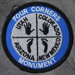 May include: Embroidered patch with a blue border and the words "FOUR CORNERS MONUMENT" in white. The center features a gray cross with black handprints and footprints, and the names of four states: Utah, Colorado, Arizona, and New Mexico.