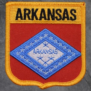 May include: Embroidered patch featuring the word "ARKANSAS" in black on a yellow border. The patch has a red background with a blue diamond containing the word "ARKANSAS" in white. The diamond is outlined in blue.